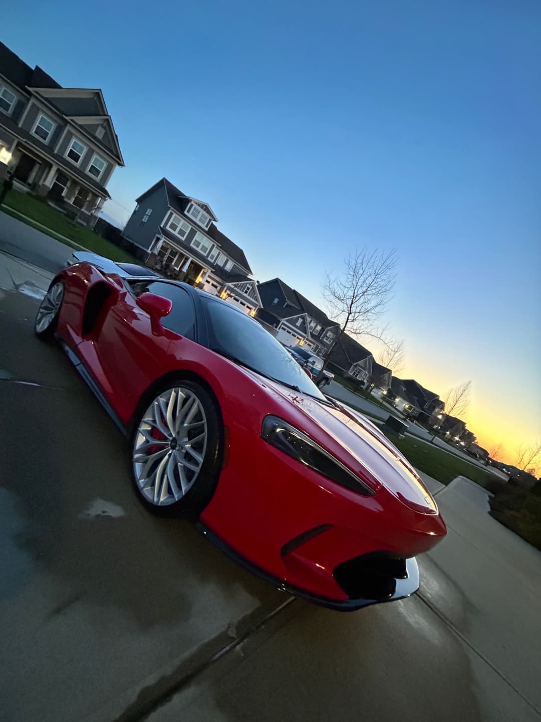 Red sports car parked in a suburban driveway at sunset, showcasing sleek design and wheels.